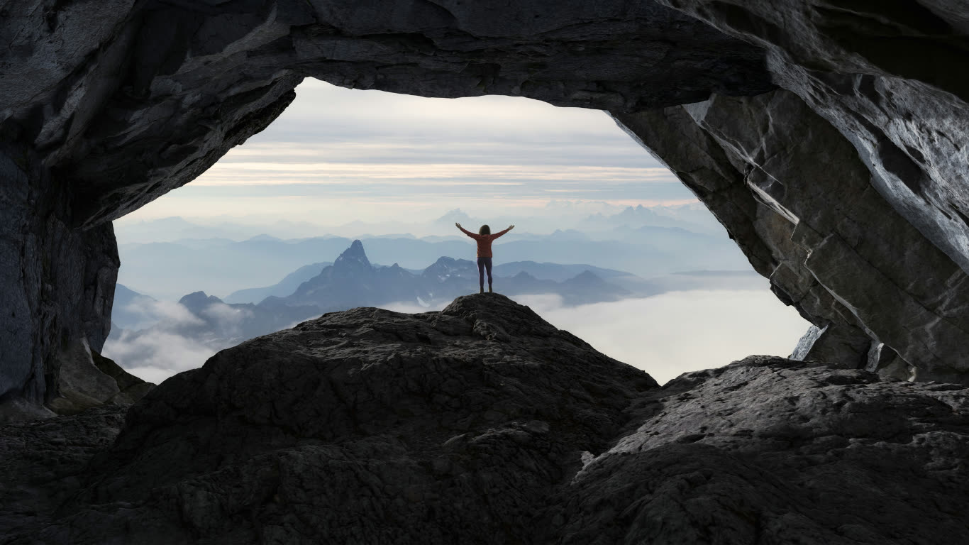 Hiker emerging from a cave overlook with arms raised at the summit